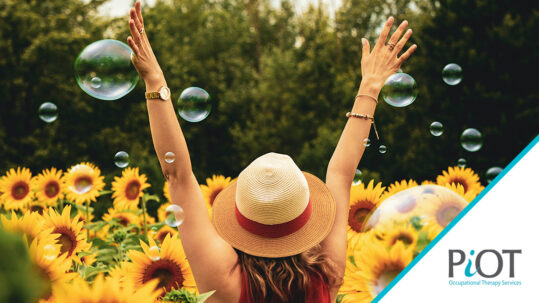 Woman wearing a sunhat rejoicing with hands in air, facing a field of sunflowers with bubbles surrounding her