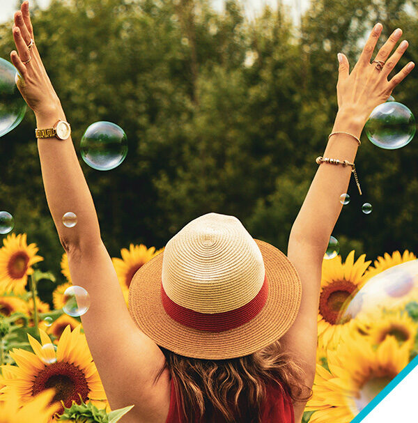 Woman wearing a sunhat rejoicing with hands in air, facing a field of sunflowers with bubbles surrounding her